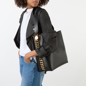 Model carrying Stella McCartney Frayme Chain Tote Bag in black with gold chain detail, styled with white tee, black jacket and blue jeans