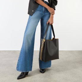 Model carrying Stella McCartney Frayme Chain Tote Bag in black with gold chain detail, styled with wide-leg blue jeans and black heeled boots