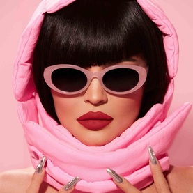 Model with KYLIE BY KYLIE JENNER Pressed Blush Powder, wearing pink oval sunglasses and pink hooded puffer wrap, bold red lip