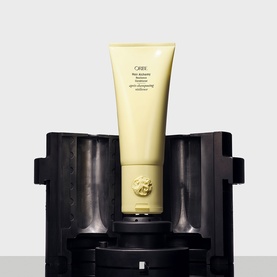 Oribe Hair Alchemy Resilience Conditioner in pale yellow tube, displayed upright on a dark decorative stand with gold emblem detail.