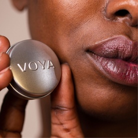 Model with Voya Balmelicious Peppermint & Vanilla Organic Lip Balm, holding silver compact tin to cheek, wearing berry-toned lip colour
