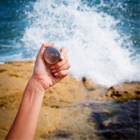 Voya Balmelicious Peppermint & Vanilla Organic Lip Balm compact tin held up against rocky coastline with ocean waves