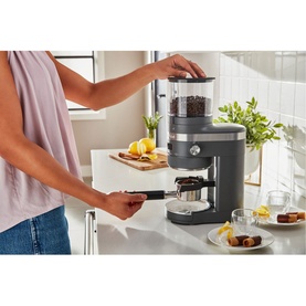 KitchenAid Artisan Coffee Grinder in grey, shown in use on a kitchen counter with a portafilter being filled with ground coffee