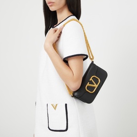 Model carrying Valentino V Logo Chain Small Shoulder Bag in black leather with gold chain strap and VLogo hardware, styled with white mini dress
