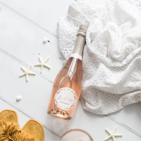 Mirabeau La Folie Sparkling Ros&eacute; bottle laid flat on white surface, styled with starfish, white lace fabric and gold sandals