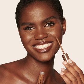 Model with Charlotte Tilbury Beautiful Skin Radiant Concealer, applying wand applicator under eye, warm deep shade finish.