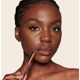 Model with Charlotte Tilbury Beautiful Skin Radiant Concealer held near lips, showcasing a medium-deep shade on deep skin tone
