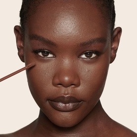 Model wearing Charlotte Tilbury Beautiful Skin Radiant Concealer, shown being applied under eye with a brush on deep skin tone