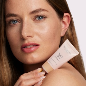 Model holding Sculpted by Aimee Tint & Glow Skin Enhancer tinted moisturiser tube in a light nude shade.