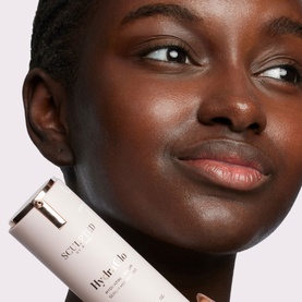 Model with Sculpted by Aimee HydraGlo Serum held against cheek, white cylindrical tube with rose gold cap