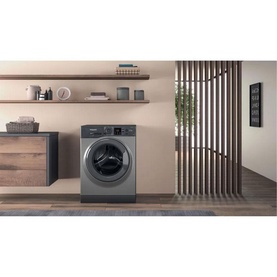 Hotpoint 8kg Freestanding Washing Machine in graphite grey, shown in a modern laundry room setting with wooden shelving and cabinetry