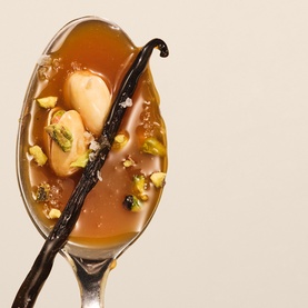 Sol De Janeiro Brazilian Joia Shampoo ingredient shot showing caramel-toned liquid on a spoon with vanilla pod, nuts and crushed pistachios