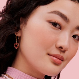 Model with Benefit Bronze Blush in Peach, wearing gold heart drop earrings and a pink top with gold chain necklace