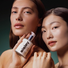 Two models with glowing skin, one holding Sisley Ecological Compound 125ml serum bottle, against a dark green background