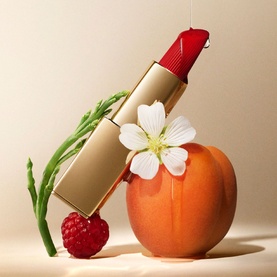 Est&eacute;e Lauder Pure Color Cr&egrave;me Lipstick in Strut, red shade in gold bullet, styled with peach, raspberry and white flower