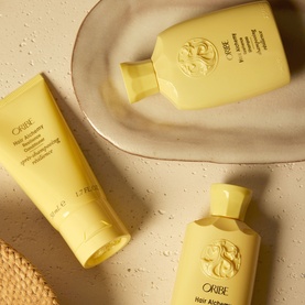 Oribe Hair Alchemy Shampoo, conditioner and travel shampoo in yellow packaging, arranged on a cream ceramic tray
