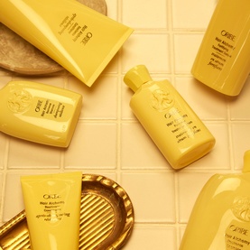 Oribe Hair Alchemy travel-size shampoo in yellow packaging, styled with matching haircare products on a cream tiled surface with gold tray.