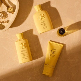 Oribe Hair Alchemy travel-size haircare set in gold packaging, including shampoo, conditioner, and treatment tube, styled on a warm beige surface.