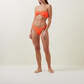 Model wearing Hunza G Jean Bandeau Two-Piece Bikini Set in orange crinkled seersucker, with ruched bandeau top and high-cut cheeky bottoms