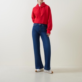 Model wearing Ganni Isoli Oversized Cropped Sweatshirt in red with drawstring hood, styled with dark blue wide-leg jeans and white trainers