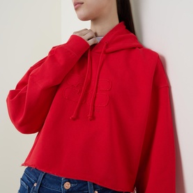 Model wearing Ganni Isoli Oversized Cropped Sweatshirt in red, hooded with drawstring and tonal logo, styled with blue denim jeans