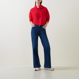 Model wearing Ganni Isoli Oversized Cropped Sweatshirt in red with blue wide-leg jeans and white flat shoes