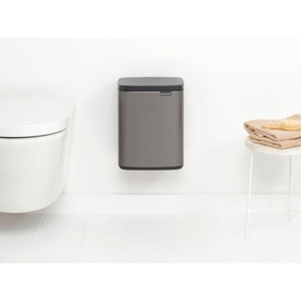 Brabantia Bo Waste Bin 7L in Platinum silver, wall-mounted in a bathroom beside a toilet, with storage basket nearby
