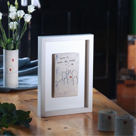 Sarah McKenna Home Is Where The Heart Is Wall Art, ceramic plaque in white wooden shadow box frame, displayed on wooden surface
