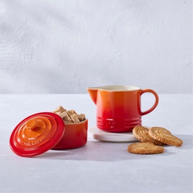 Le Creuset Stoneware Milk & Sugar Set in orange, lidded sugar pot with brown sugar cubes and creamer jug, styled with biscuits
