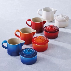 Le Creuset Stoneware Milk & Sugar Set in orange, shown alongside matching sets in red, blue, and white stoneware