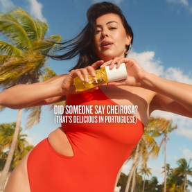 Model wearing red cutout swimsuit, holding Sol De Janeiro Rio Radiance Perfume Mist, posed against tropical palm tree backdrop.