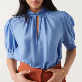 Model wearing Bash Tyler Top in blue, featuring short balloon sleeves, stand-up collar and keyhole neckline, styled with rust trousers