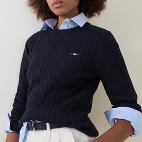 Model wearing Gant Cable Knit Logo Sweater in dark blue with crew neck, styled over a light blue shirt with white trousers and black belt