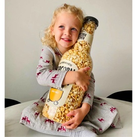 Child hugging Popcorn Kitchen Simply Sweet Giant Monet Bottle 550g, a large clear popcorn-filled gift bottle with cream and blue label
