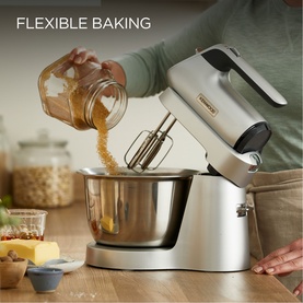 Silver Kenwood stand mixer with stainless steel bowl, ingredients being poured in, styled on a kitchen worktop with baking items nearby.