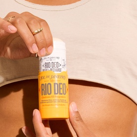 Model with Sol De Janeiro Rio Deo 62 deodorant stick held against bare midriff, yellow packaging with branded label.