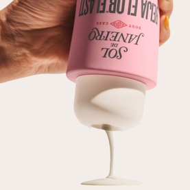 Sol De Janeiro Beija Flor Body Wash pink bottle held upside down, dispensing white creamy formula into a stemmed glass bowl