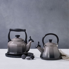 Le Creuset Kone Kettle With Fixed Whistle 1.6L in grey enamel, shown in two sizes with black handles on a neutral background