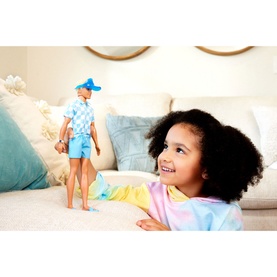 Young girl smiling and holding Barbie Blonde Ken Doll dressed in blue check shirt, shorts and blue hat with beach accessories