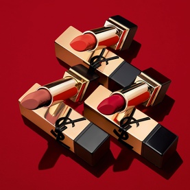 YSL Rouge Pur Couture Lipsticks in assorted red and nude shades, gold cases with black YSL logo, arranged in a stack on red background