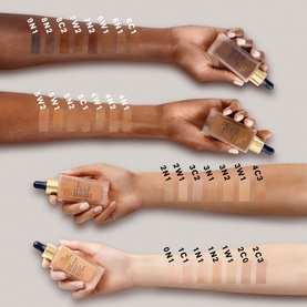Four arms showing Est&eacute;e Lauder Futurist SkinTint Serum Foundation SPF 20 swatches across multiple shades, with bottles held alongside
