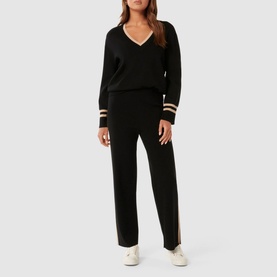 Model wearing Forever New Hattie Knit Sweater in black with V-neckline and contrast gold stripe trims, styled with matching black knit trousers
