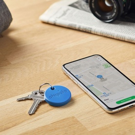 Chipolo ONE Bluetooth Item Finder in blue, disc-shaped tracker on a keyring beside a smartphone displaying a map location app