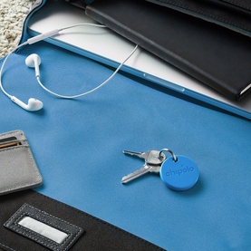 Chipolo ONE Bluetooth Item Finder in blue, attached to keys on a desk beside earphones, wallet, and laptop case