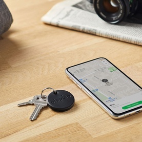 Chipolo ONE Bluetooth Item Finder in black, attached to keys on a wooden desk beside a smartphone displaying a map tracking screen