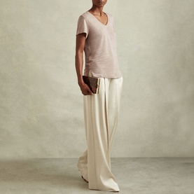 Model wearing Reiss Lottie Marled Solid T-Shirt in pink linen with V-neckline, styled with wide-leg cream trousers and a small clutch bag