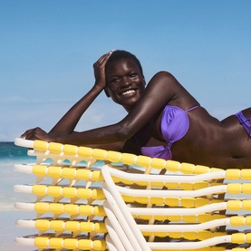Sol De Janeiro Delicia Drench Body Butter lifestyle shot featuring a smiling model relaxing on a yellow and white beach lounger by the sea.