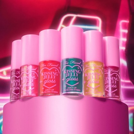 Too Faced Kissing Jelly Lip Oil Gloss collection in assorted shades displayed on a tiered pink stand with neon backdrop