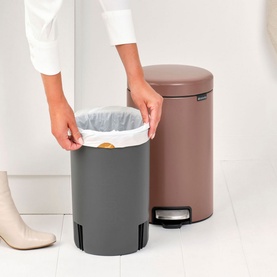 Brabantia NewIcon Pedal Bin 12L in Taupe beside a grey bin, model removing liner bag. Chrome pedal and rounded lid visible