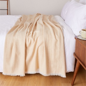 Foxford Extra Large Lambswool Throw in camel, herringbone weave with self-fringed trim, draped over a white bed